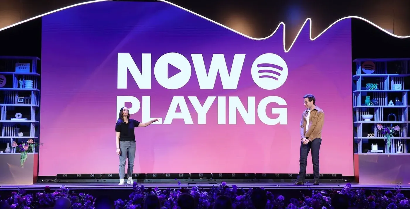 Two presenters stand onstage before a giant purple screen reading "NOW PLAYING" with the Spotify logo.